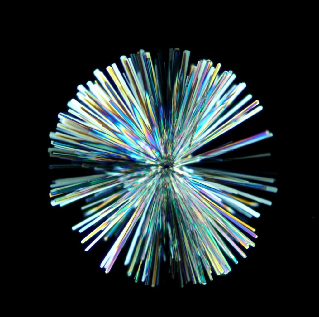 Iridescent rod-shaped crystals extending out of a single point to form a ball on a black background. 