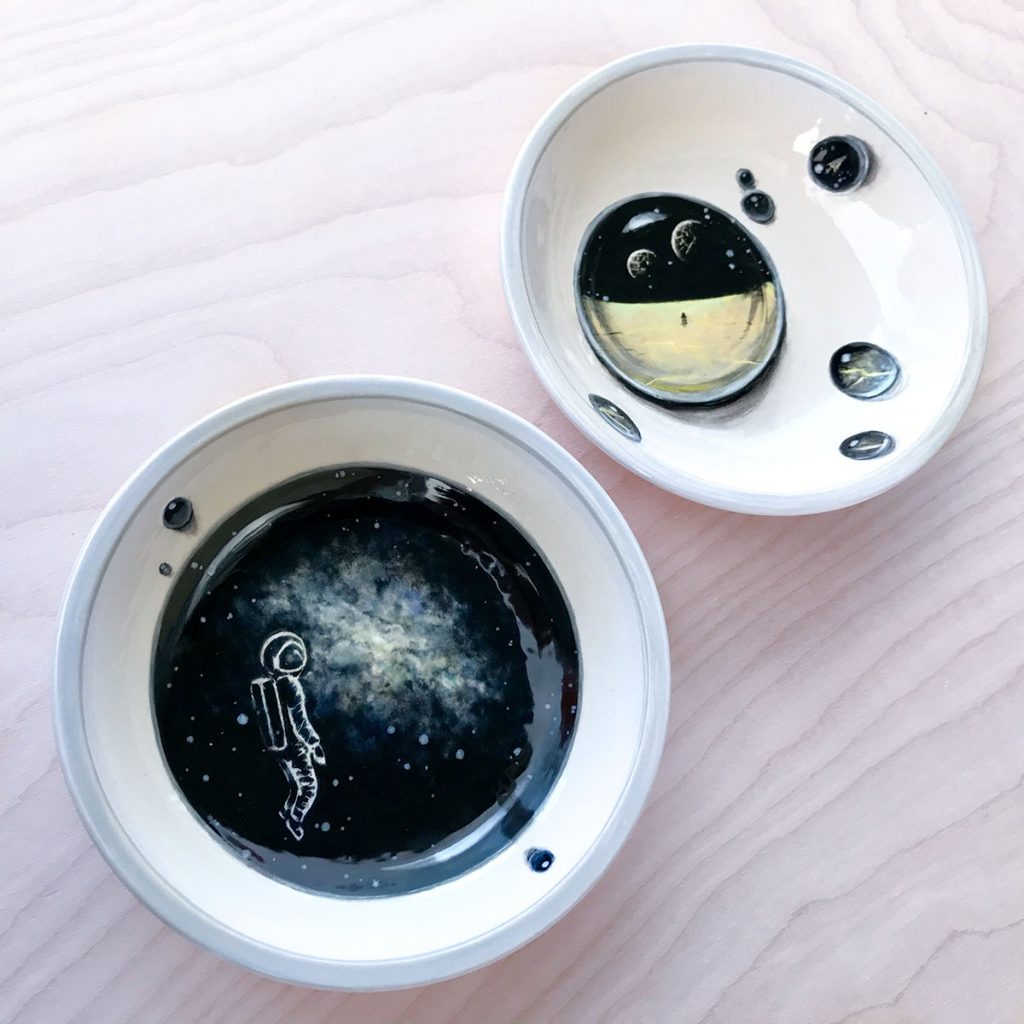One bowl has the white outline of an astronaut floating in space in front of a white-blue nebula, and the other bowl has droplets painted on it. Within the droplets are paintings of another bowl, "The Invasion."