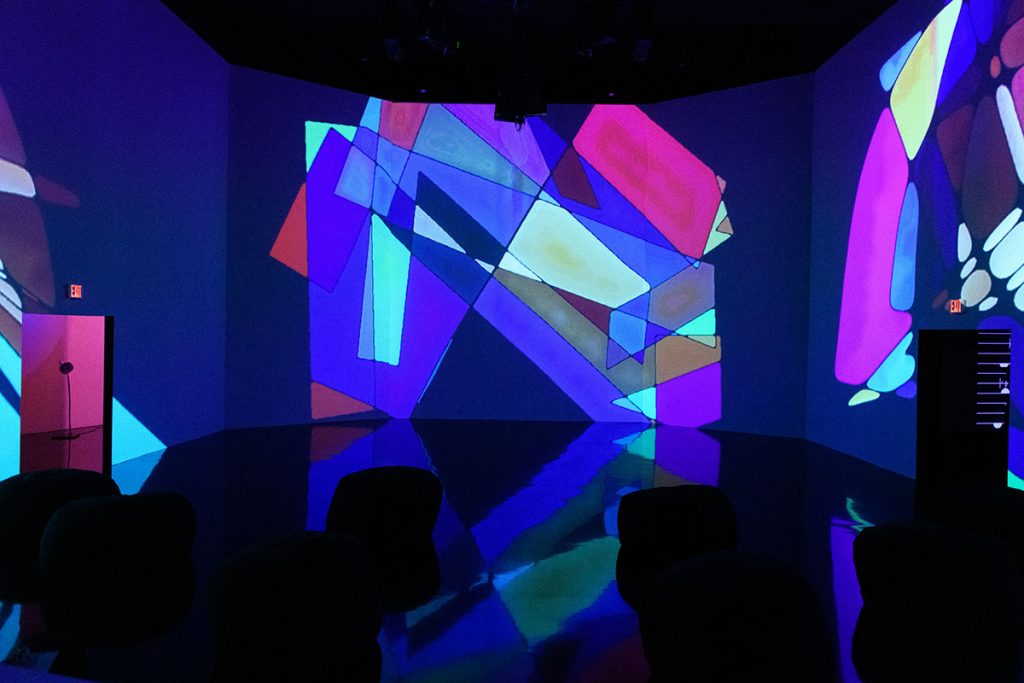 Colorful, geometric shapes are projected onto three large, open white walls in the dark exhibition space. 