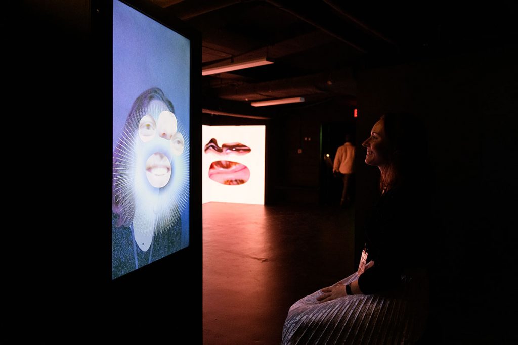 Woman sitting in front of a display, with her face on the screen. Her eyes, mouth, and nose are replaced with enlarged versions of someone else's eyes, mouth, and nose. 