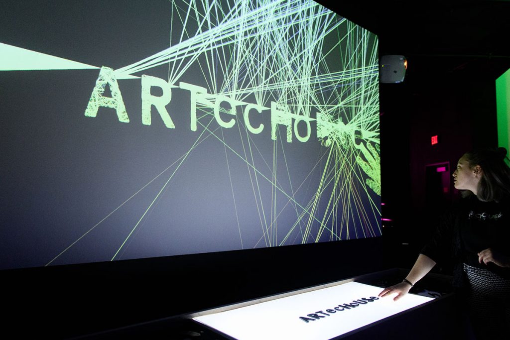 Woman places plastic letters spelling "Artechouse" on a backlit table. The letters are displayed on a screen with yellow lines bouncing off them. 