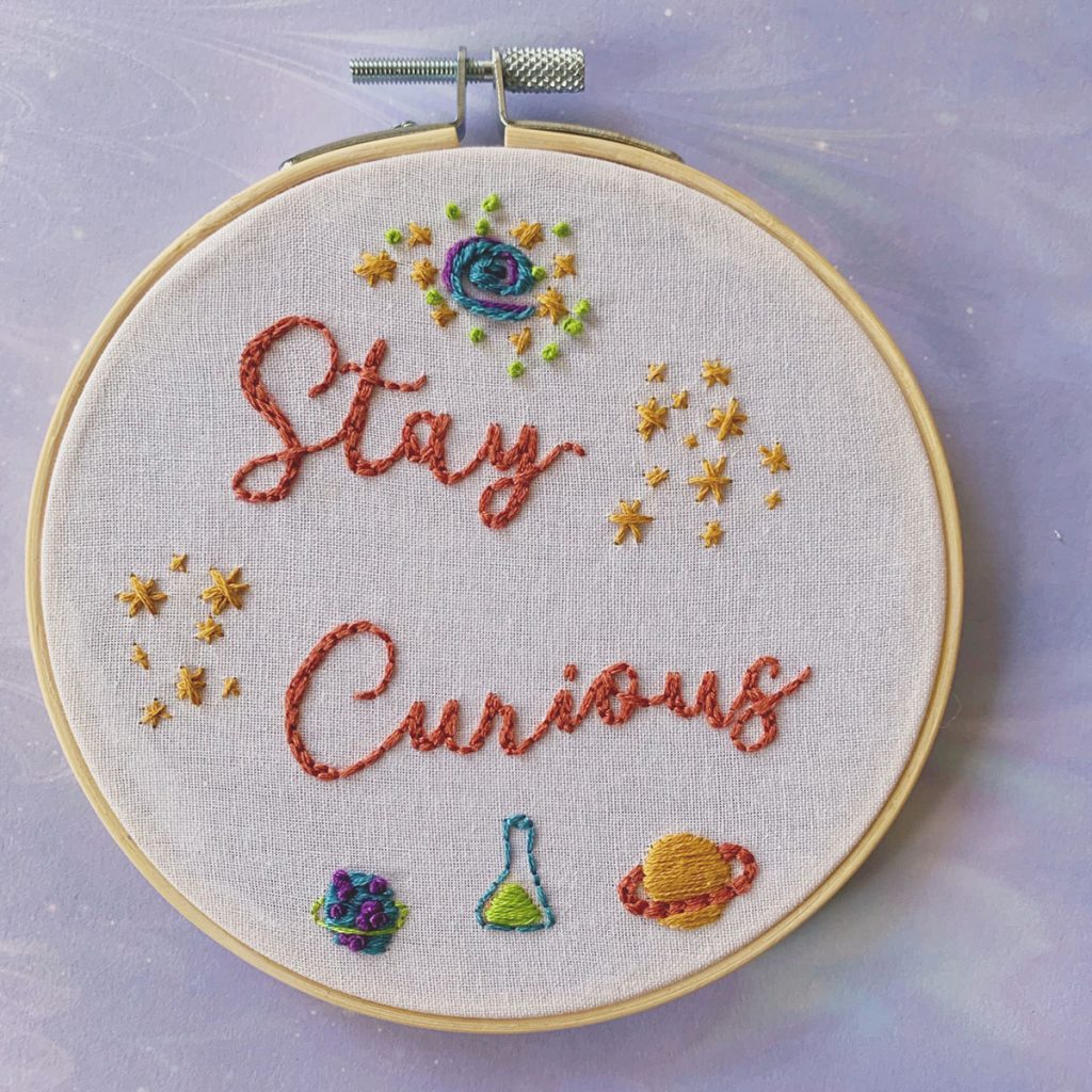 Embroidery piece that says "Stay Curious" surrounded by stars, a nebula, planets, and an Erlenmeyer flask. 