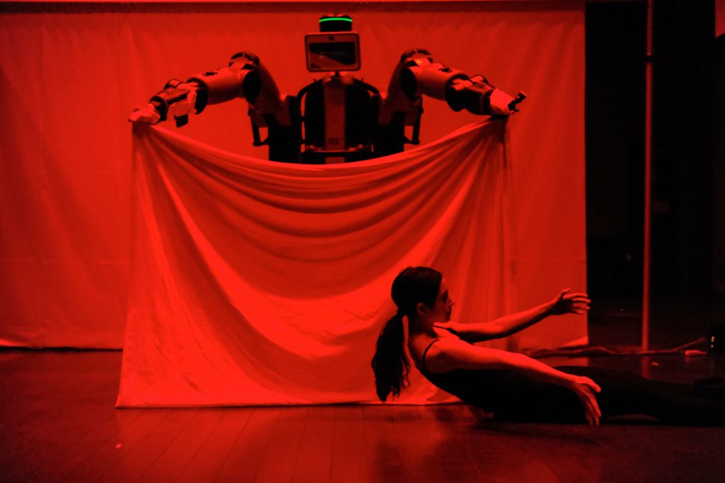 A large robot holds up a white sheet while Catie dances on the floor in front of the sheet. 