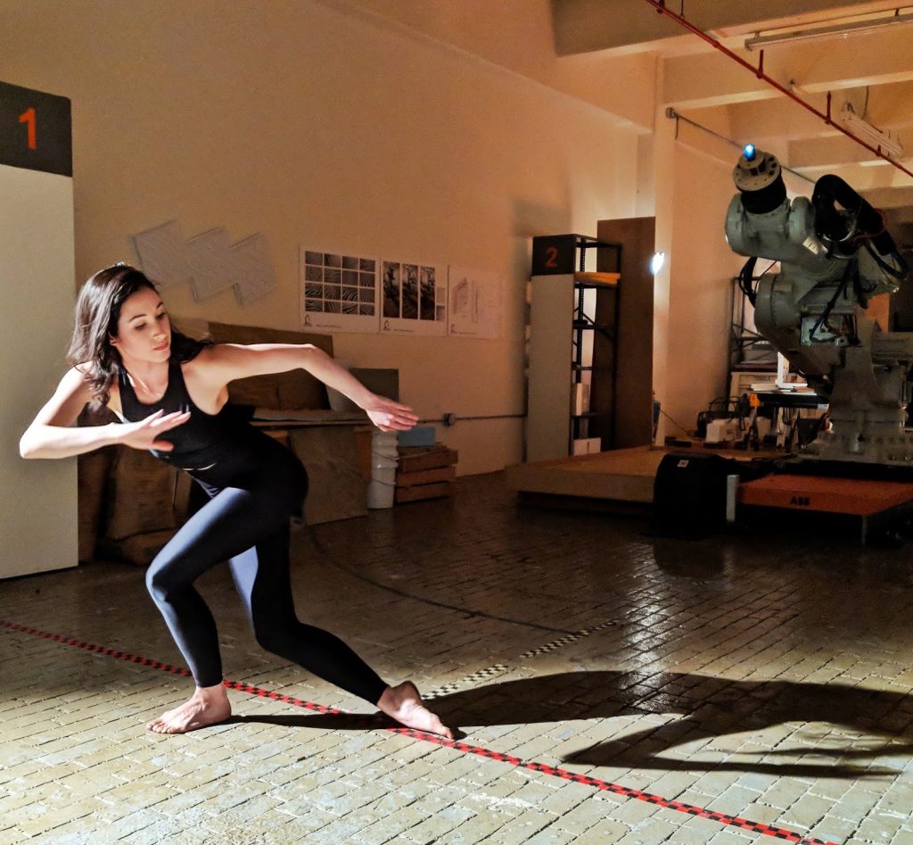 Catie posing in front of a large robot that is mimicking her posture. 