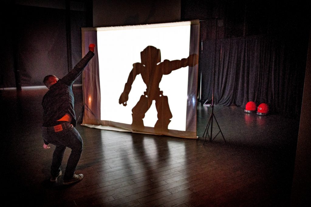 Participant posing in front of an illuminated screen with their arm out. Behind the screen, you can see the silhouette of a robot mimicking the participant's pose.