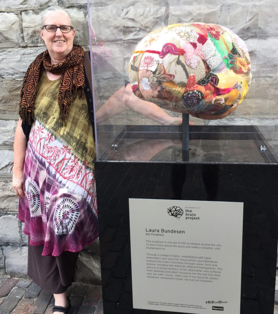 Laura Bundesen standing next to her brain sculpture entitled Not Forgotten. It is a three dimensional structure of a brain made with different fabrics and other materials. 