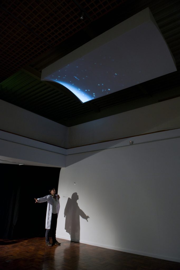 Woman stands beneath a panel mounted on the ceiling. The panel has blue micro-algae, which looks like fuzzy dots, projected onto it.