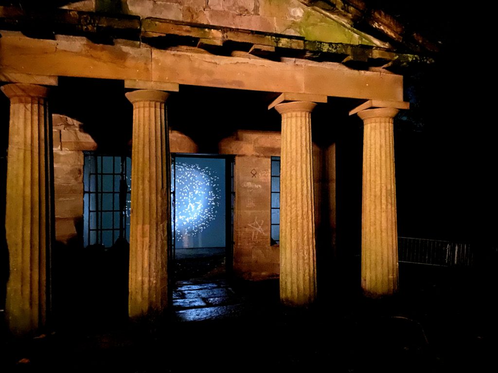 Video still of a glowing orb emitting light and sparkles. Video is displayed on the entrance of a building that looks like an ancient Greek building. 