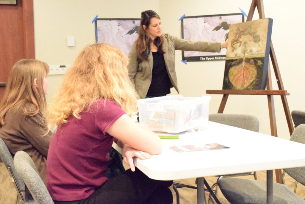 Annette Lee discussing a painting in front of 2 girls sitting at a table. 