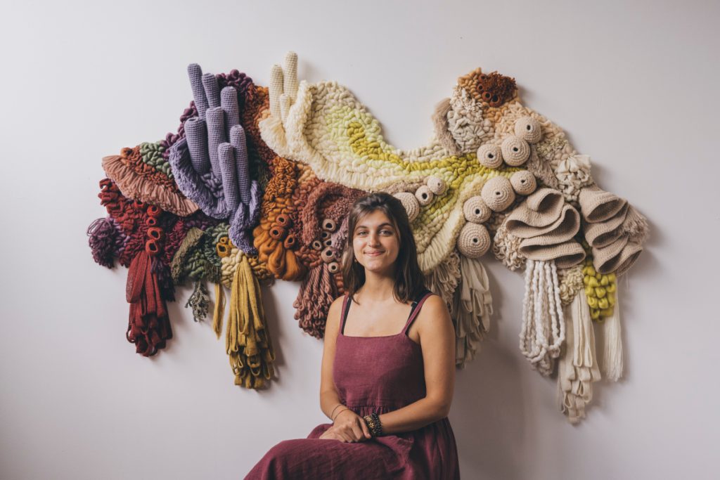 Vanessa sitting and smiling in front of one of her tapestries. 