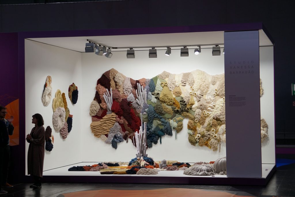 Multiple tapestries, some on the ground, some on the wall, and a large one hanging in the back within a large display. There are many colorful corals, but they become white toward the right side of the display. 