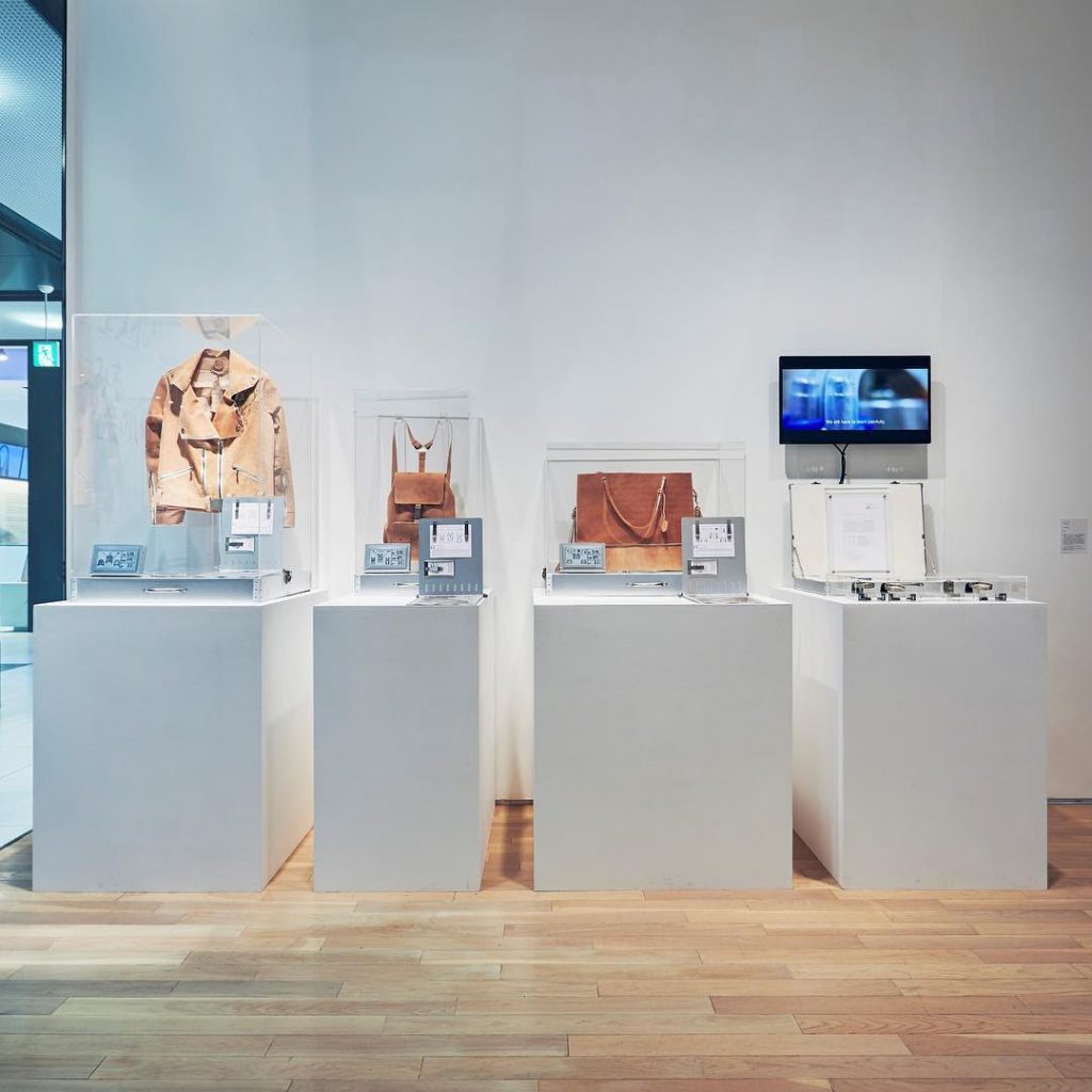 A gallery display of Pure Human 1 with a leather jacket, two bags, and a briefcase containing a patent placed on white podiums. 