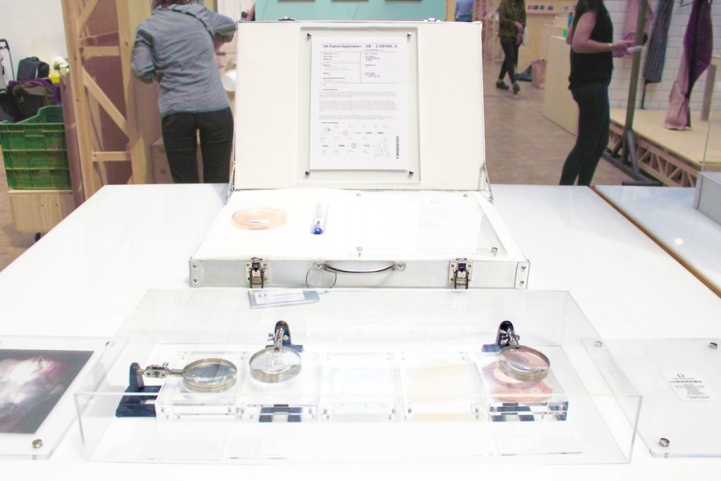 A gallery display of Pure Human 2 with a briefcase containing reagents and a patent on a table. The table also contains a glass case with magnifying glasses hovering over pieces of leather. 