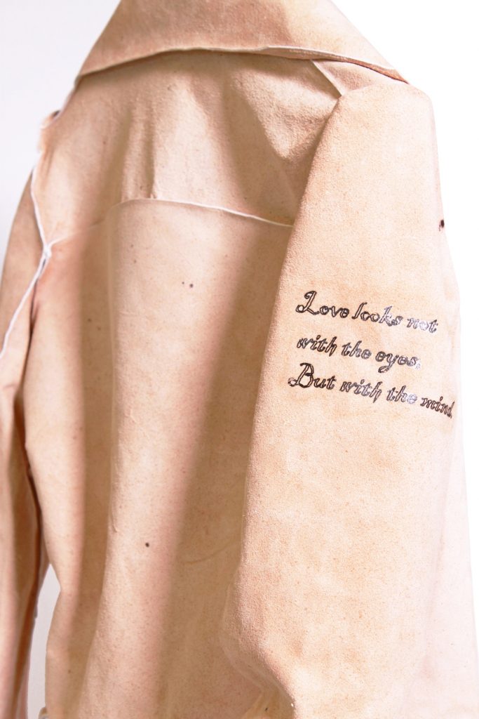 A pale peach-colored leather jacket seen from behind with a tattoo on the right sleeve saying "Love looks not with the eyes, But with the mind."