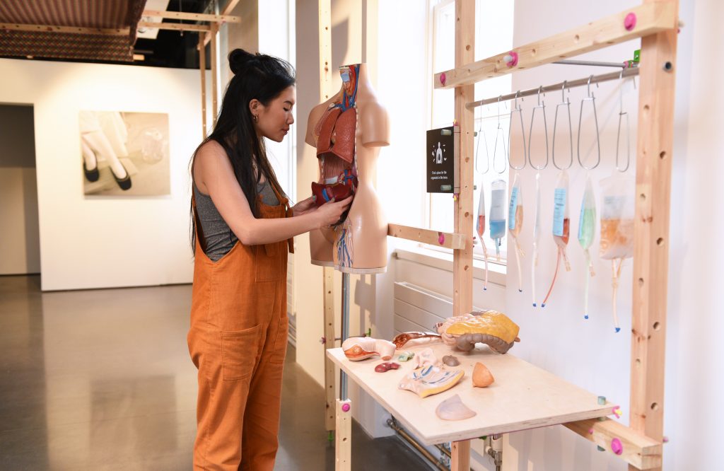 A gallery display of The Self-Donor Workshop with a women replacing an organ from a model of a human torso. Artificial organs and colorful IV bags are on a table nearby. 