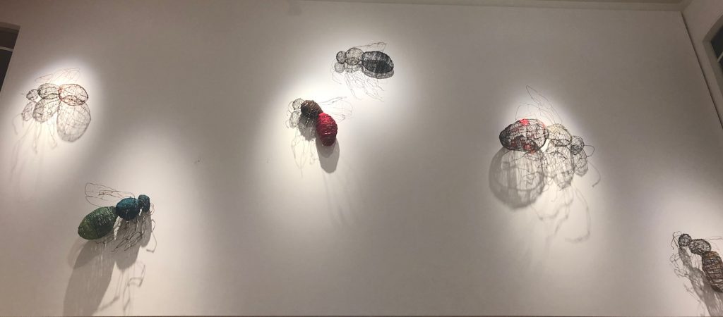 Large bees made of wire hanging on a white wall. A spotlight shines on each bee to create shadows on the wall. 