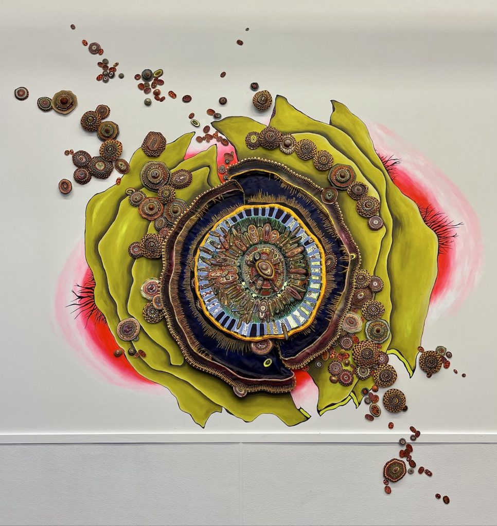 Before the Split (2020) by Amie Esslinger. A large layered ovoid-shaped cellular body on the verge of splitting in two, where the position of the two halves suggests they are separating by pulling diagonally away from each other. A small central oval, like an eye, is surrounded by membranous layers of vibrant patterns and other similarly sized ovals resembling cellular organelles. The centre is mostly green, with the smallish ovals arranged in a ring. Some of these ovals cross over to the next blue outer ring punctured with pores through which the small ovals appear to move through. The next outer ring is yellow, followed by a thicker membrane of blue, then a layer of mixed green and pink. Beyond that are several layers of green yellow where several small and tiny oval- and circle-shaped cellular bodies appear to come out of the darker shaded central body. The split is clearest in the green-yellow layers at the northwest and southeast points of the whole work, where there are cracks. Behind the work is a pool of red with some black branching structures extending outward.