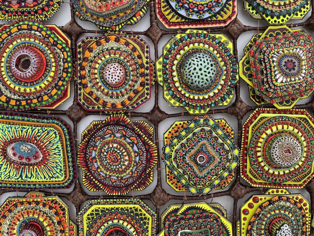 A close-up of Cell Grid (Brown) (2019) by Amie Esslinger. A brown textile grid with 6 columns and 5 rows. Each cell in the grid is occupied by an ovoid body resembling a biological cell. These cells have various patterns and colours like green, bright yellow, blue, red, orange. Some are shaped like flowers, some are square with the corners cut off, some are combinations of both shapes, with multiple layers. The grid frame lines extend beyond the frame of the grid holding all the cells, and the bottom has long threads flowing down like a fringe (same material as the brown grid). This close-up shows 3D layering incorporated into the work.