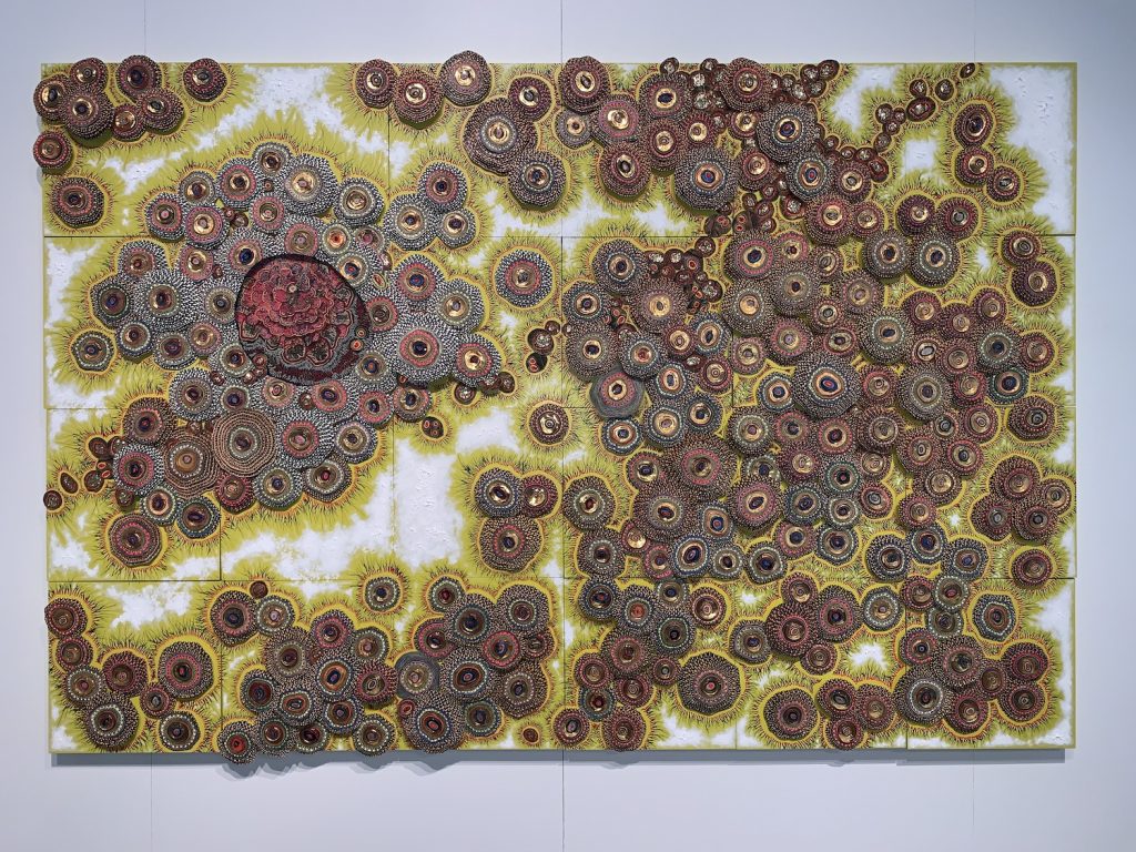 Mother and Colony (2017) by Amie Esslinger. Horizontally oriented rectangular work with large clusters of circular cell-like bodies. The clusters concentrate towards the top left around a larger circular body and towards the right side in a mass. The circular cells appear to be coloured either blue-grey or mauve, each with its own central nucleus and surrounded by kiwi green hair-like structures projecting outward. The gaps between clusters is white. The inside of the large circular body towards the left contains a layered structure that looks like an open camellia flower. 