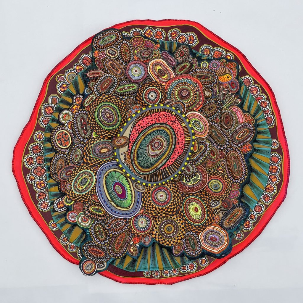 Pile Up, Pile On, It Never Ends (2021) by Amie Esslinger. A large cellular body with a bright red border. Within is a multitude of small oval or gem-like shapes to resemble smaller cells with various patterns and colours. One large oval shape, bordered by a ring of alternating yellow and blue, is centred and the size changes and reduces towards the outer edge. Beyond the ovals immediately connected to the central oval is a fringe of flat yellow spikes on blue-black material. Beyond that is a ring of gem-like shapes, similarly coloured with red-orange patterns, gem-cut white borders, and a yellowish wall. The bright red border finishes the piece.