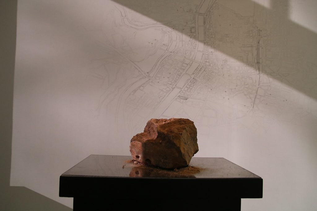 Slab of red clay on a pedestal. Behind it is a black and white illustrated map.