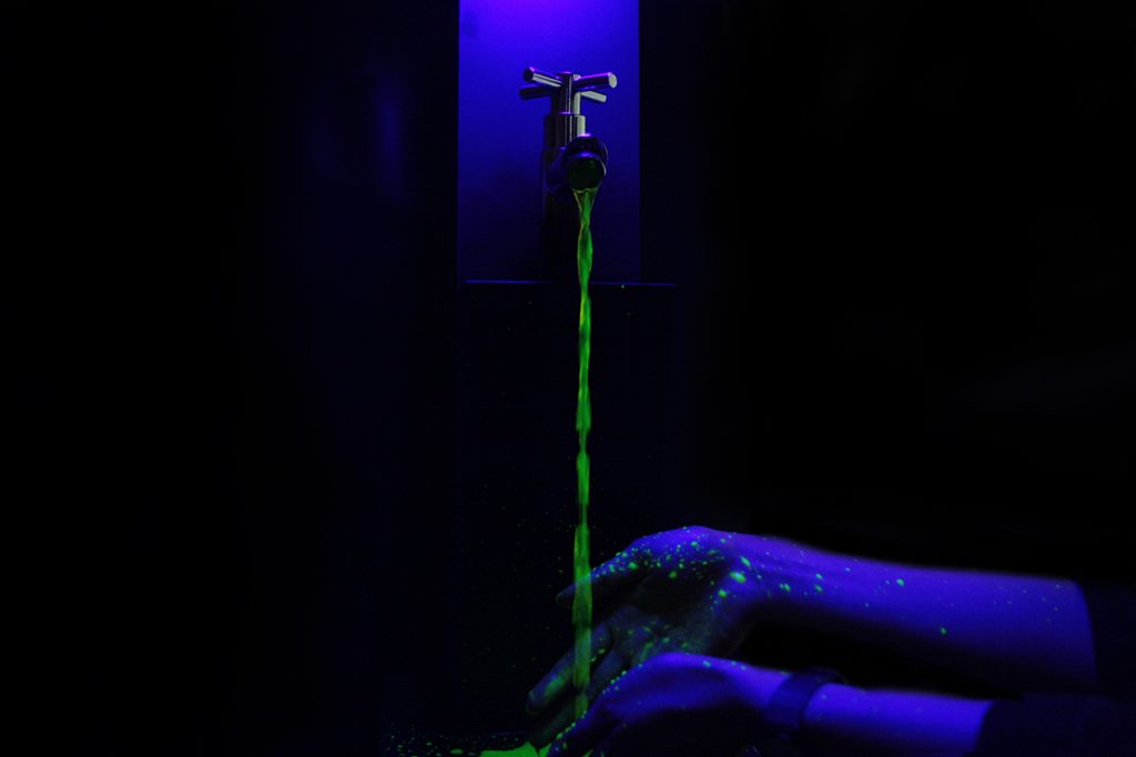 Photo of hands below a running faucet. The lighting is dark with blue lights, so the hands appear blue and the water glows fluorescent green. 