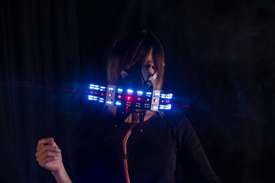 Woman stands in a dark room with a mask covering her mouth and nose. The mask contains tech and projects blue lights. 