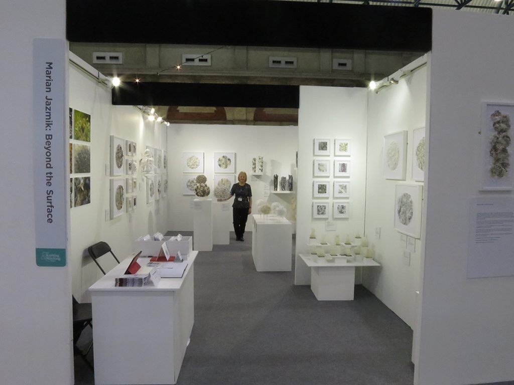 Marian Jazmik stands in the exhibition space with the walls covered by her framed works. 