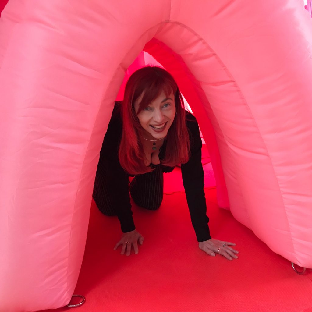 A photo of a smiling woman on her hands and knees. She is coming out of a pink inflated arch.
