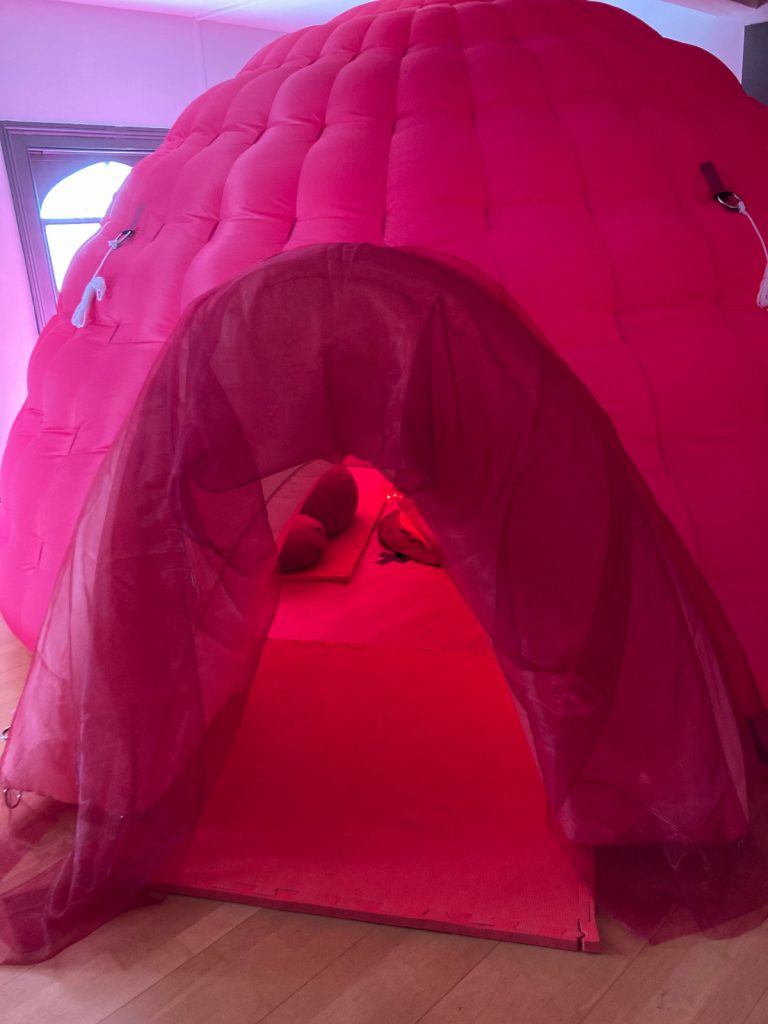 A photo of a large inflated pink dome. We can see through its entrance to a dimly lit interior.