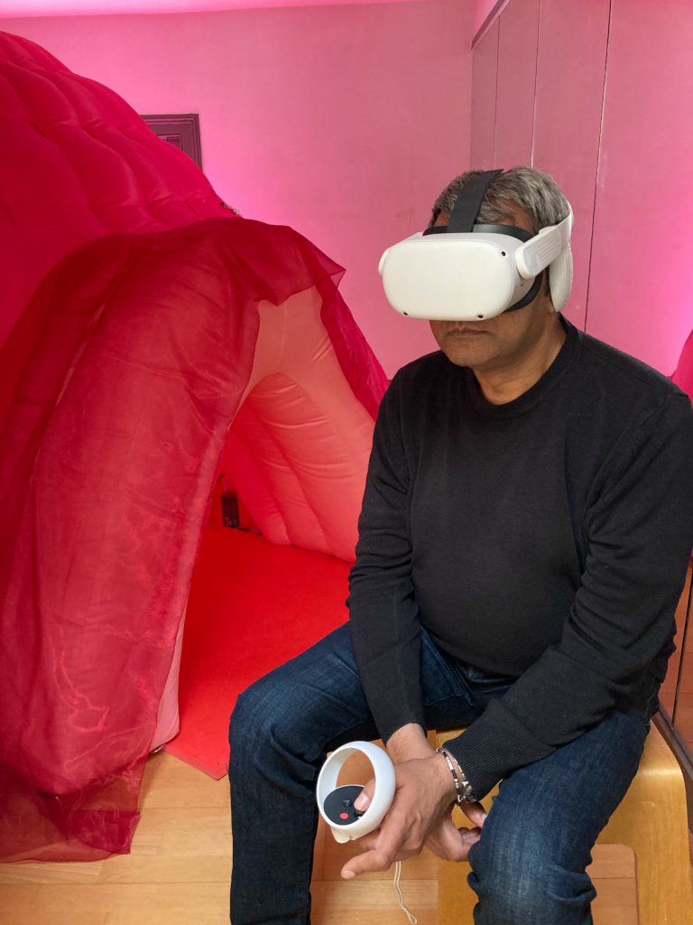 A photo of a man sitting on a stool. He wears a white headset over his eyes and holds a controller. In the background is the entrance of a large inflatable pink tent.