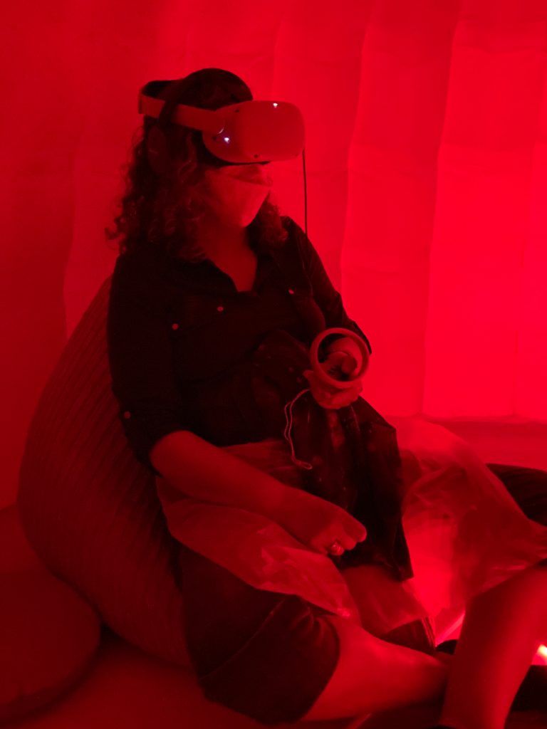 A photo of a woman wearing a headset over her eyes and headphones, sitting on a beanbag chair and holding a controller. The lighting is dim and red-coloured.