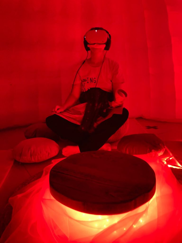 A photo of a woman wearing a headset over her eyes and headphones sits behind a small table. The space is dimly lit and red-coloured, with a gold light source beneath the table.