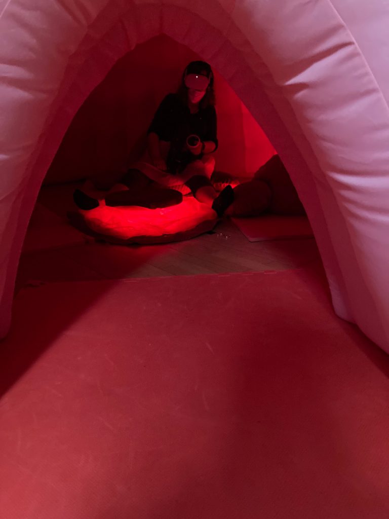A photograph of a woman wearing a headset over her eyes sitting inside a pink tent
