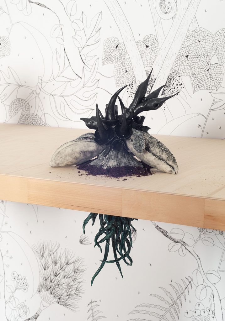A sculpture of a black spikey plant with a base that looks like lobster claws sits on top of a table. Underneath the table, it's dark green roots are visible.