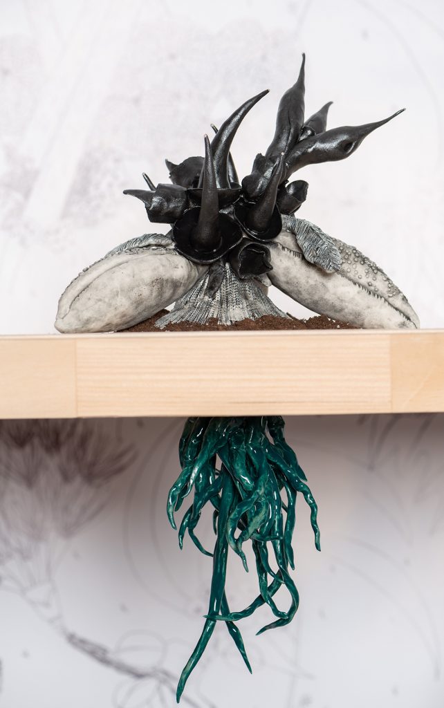 A sculpture of a black spikey plant with a base that looks like lobster claws sits on top of a table. Underneath the table, it's dark green roots are visible. 