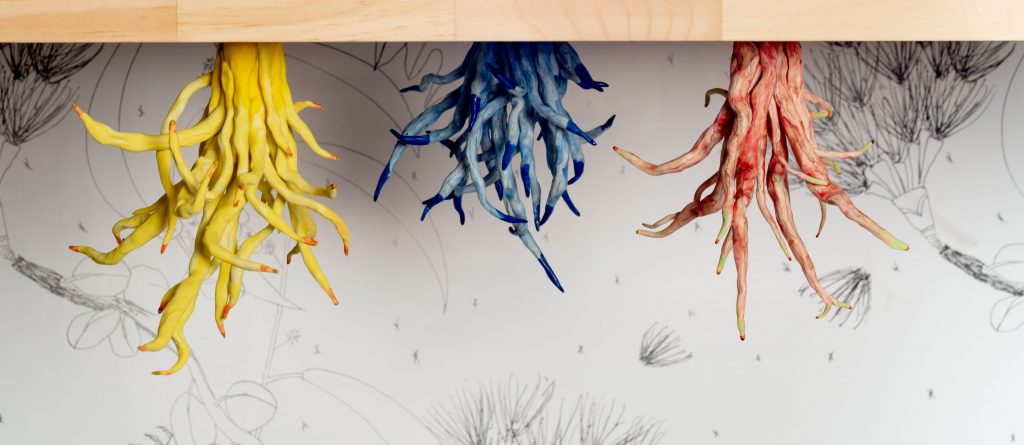 Three sets of sculpted roots -- one yellow, one blue, and one red -- emerging from below a wooden table.