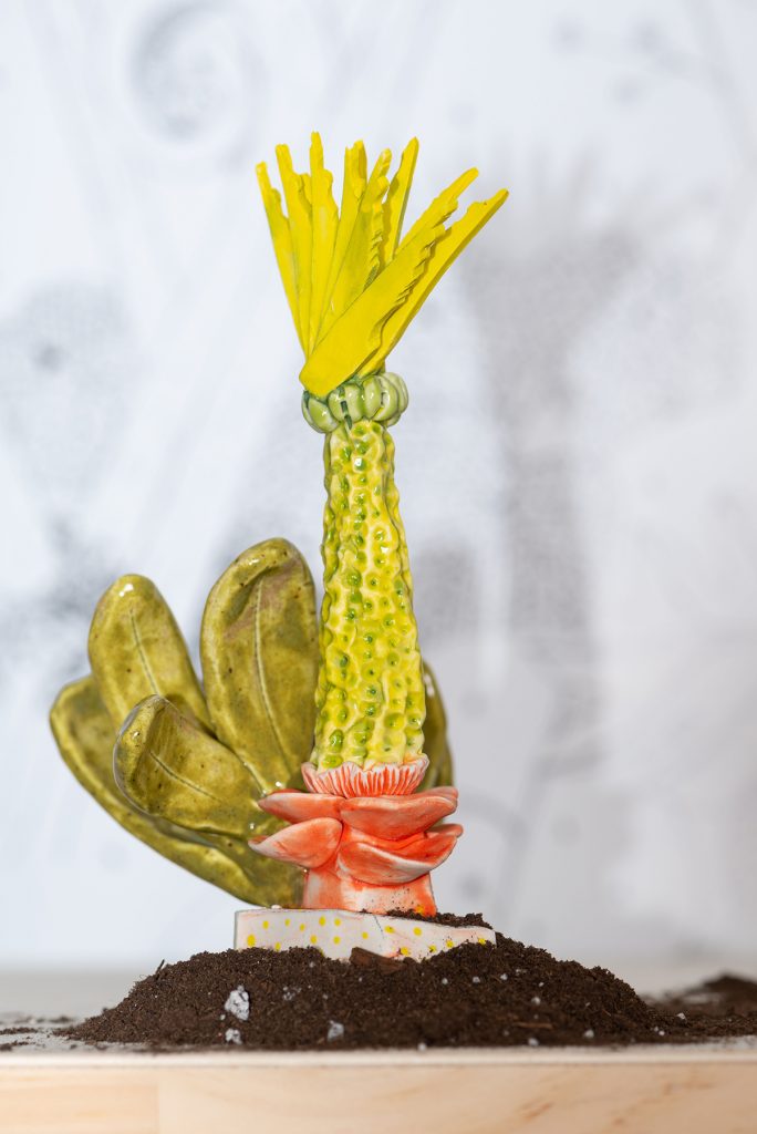 A sculpture of a yellow-green plant that emerges from a pink flower-like base. Coming from the side are large leaves. At the top of the plant are sharp yellow spikes.