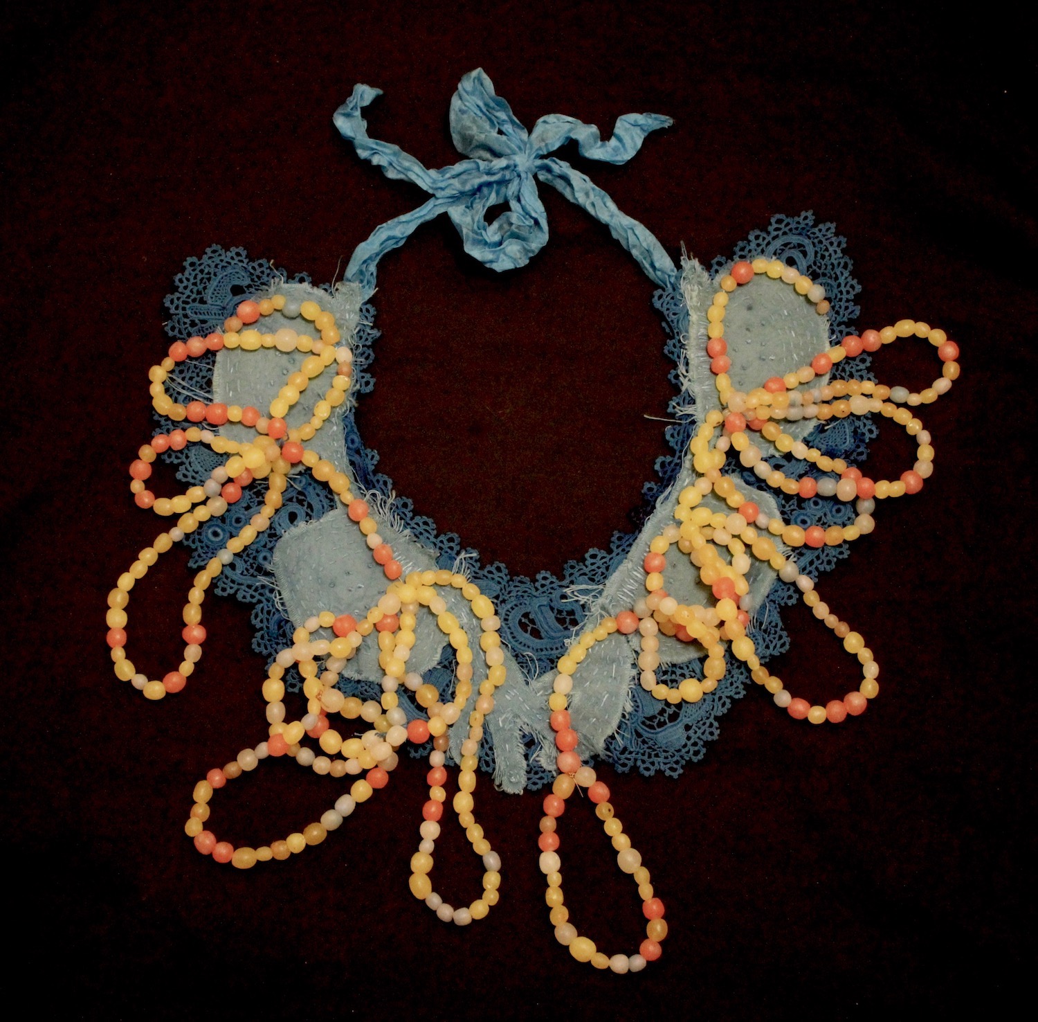 A multi-layered necklace with a flat blue lace base, grey fabric overlay, and an orange, white, and yellow bead string looped over the top.