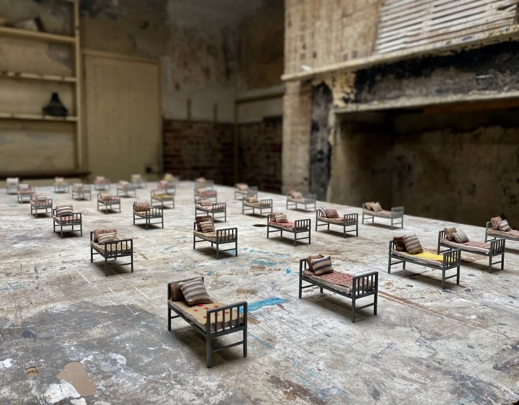 Many miniature beds with a blanket and pillow each are lined up on a worn-looking table in a room with crumbling walls.