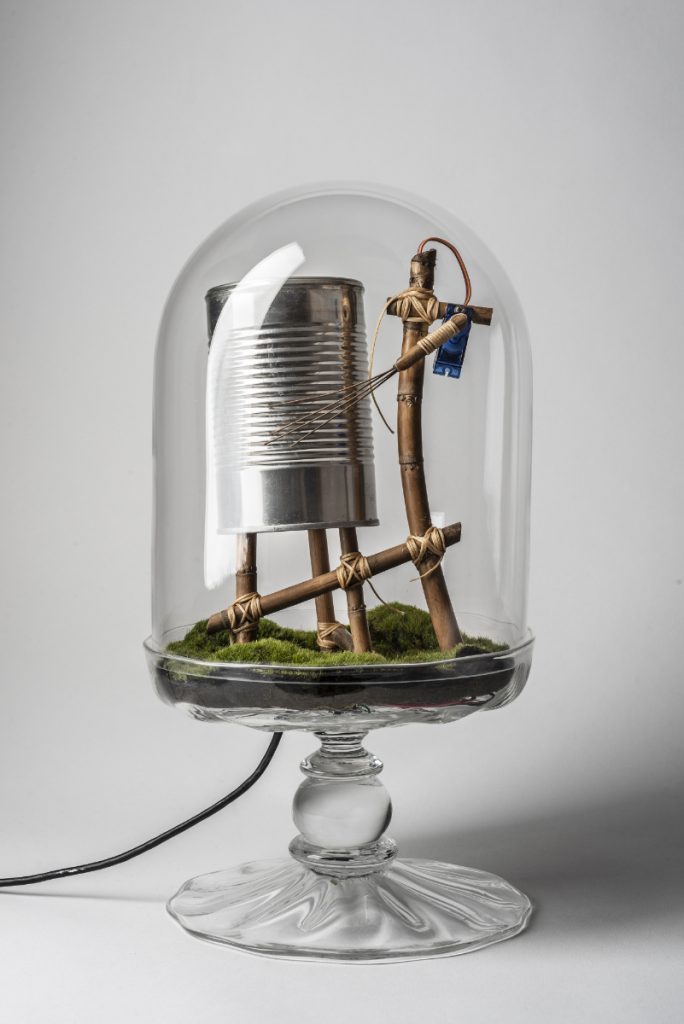 Clear glass terrarium containing a wooden mechanism that rakes metal wires across a tin can, sitting on top of moss