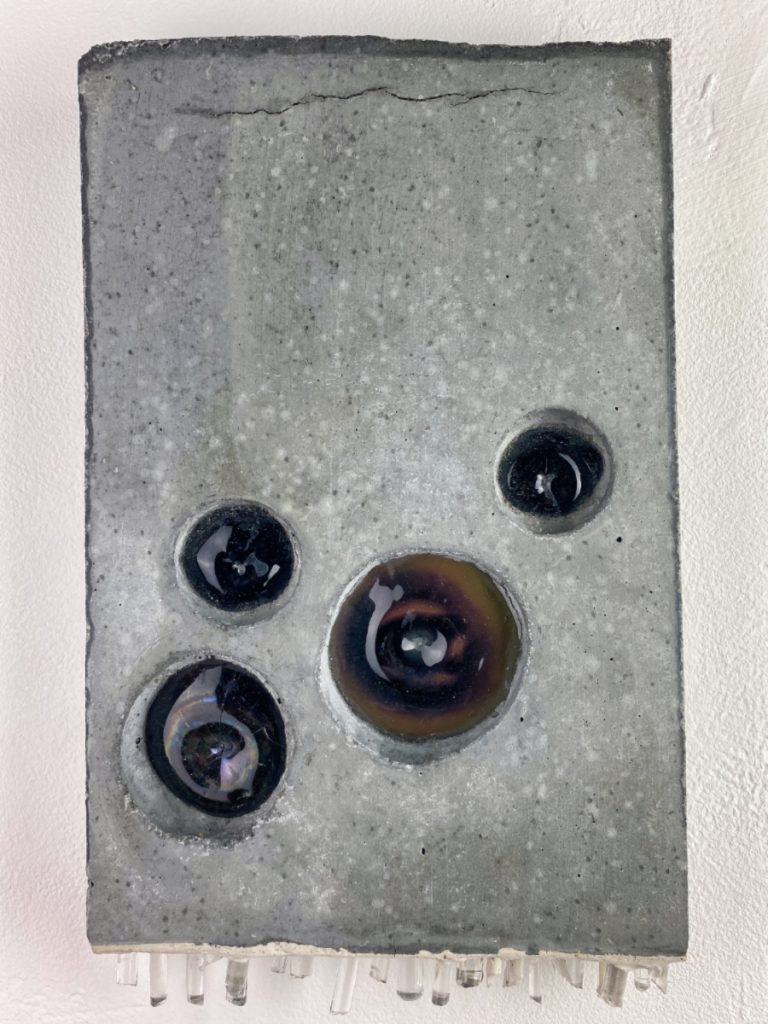 A grey rectangular slab of concrete embedded with dark blue and brown glass balls and clear crystal-like glass sticking out from the bottom