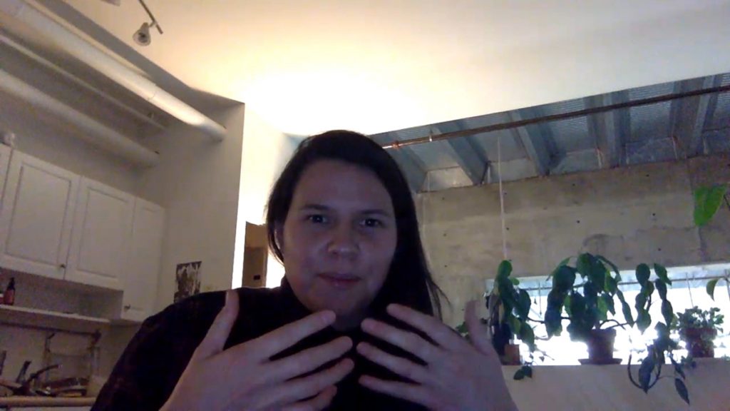 A person faces the camera and holds up their hands, showing their fingers. They look like they are in mid-speech. Plants line a window in the background.