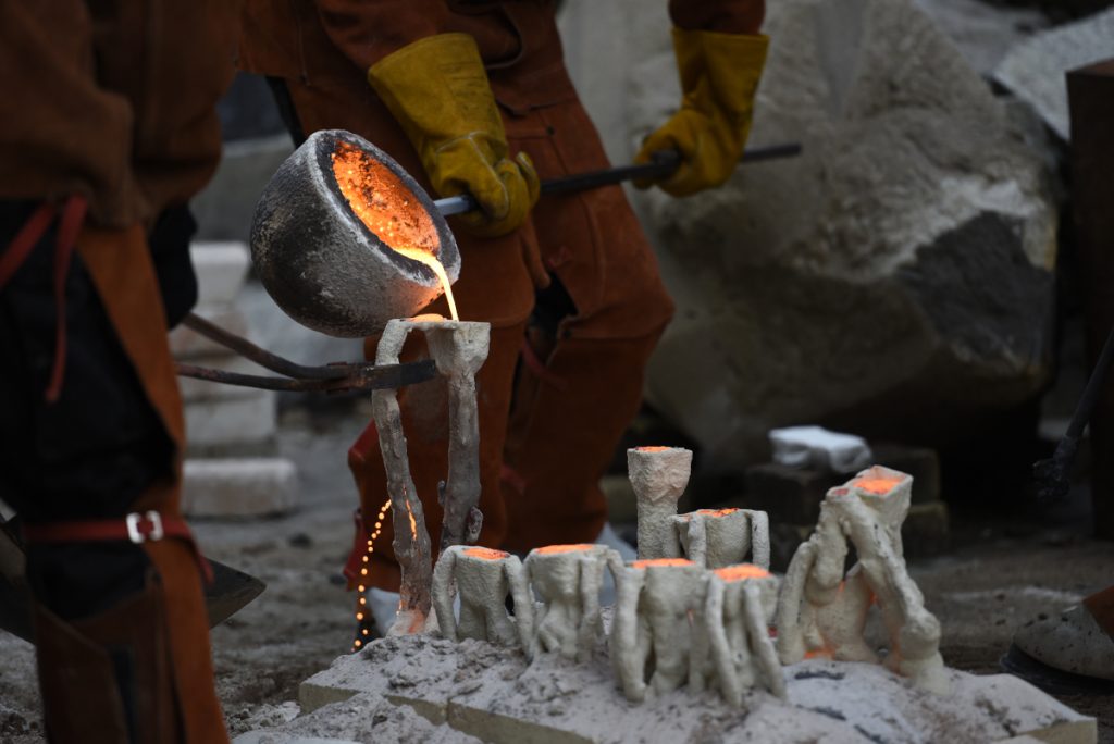 A gloved figure pouring hot iron from a large ladle into ceramic moulds.