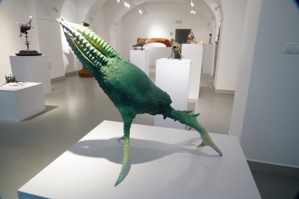 A green sculpture with branching legs and several ribbed appendages extending upwards to meet at their tips. The sculpture is on display at a gallery among other organic sculptures.
