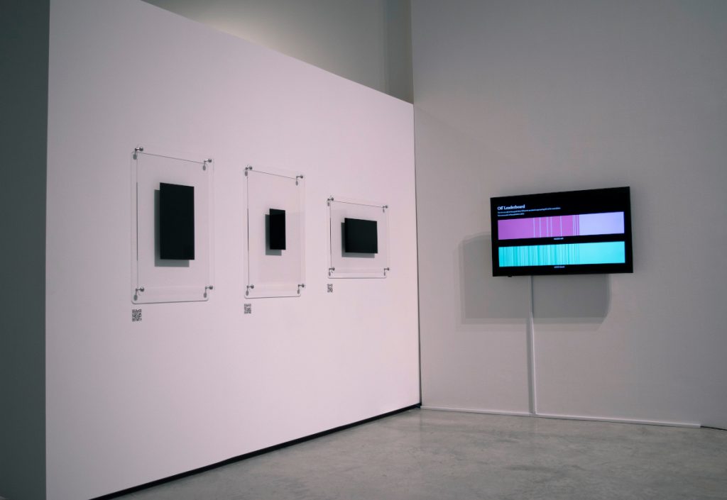 Three black rectangles displayed on glass backing on a white wall in a gallery. A screen showing two coloured bars is on the wall adjacent.