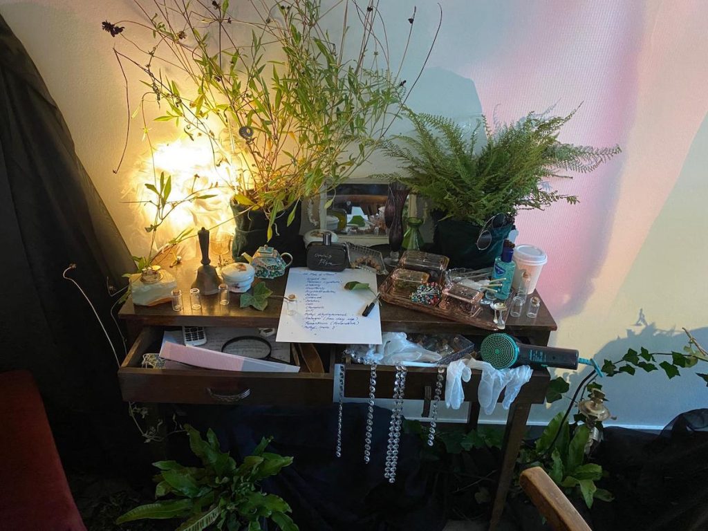 A wooden desk covered with plants and objects, such as a coffee cup, a bell, hand sanitizer, and jewelry boxes