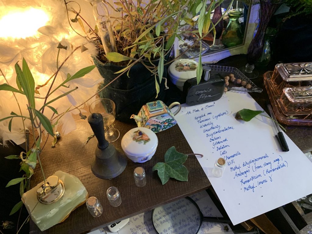 The corner of a desk covered in plants and objects, such as a bell, a small teapot, a bottle of perfume, and a piece of paper reading "All Made of Flowers, upcycled rose, narcissus (synthetic), waterlily, uncertainty, bicyclononalactone, beehives, goldenrod, isolation, cats, chamomile, WiFi, methyl dihydrojasmonate, ambergris (from clay sage), romanticism (romandolide), methyl lemone