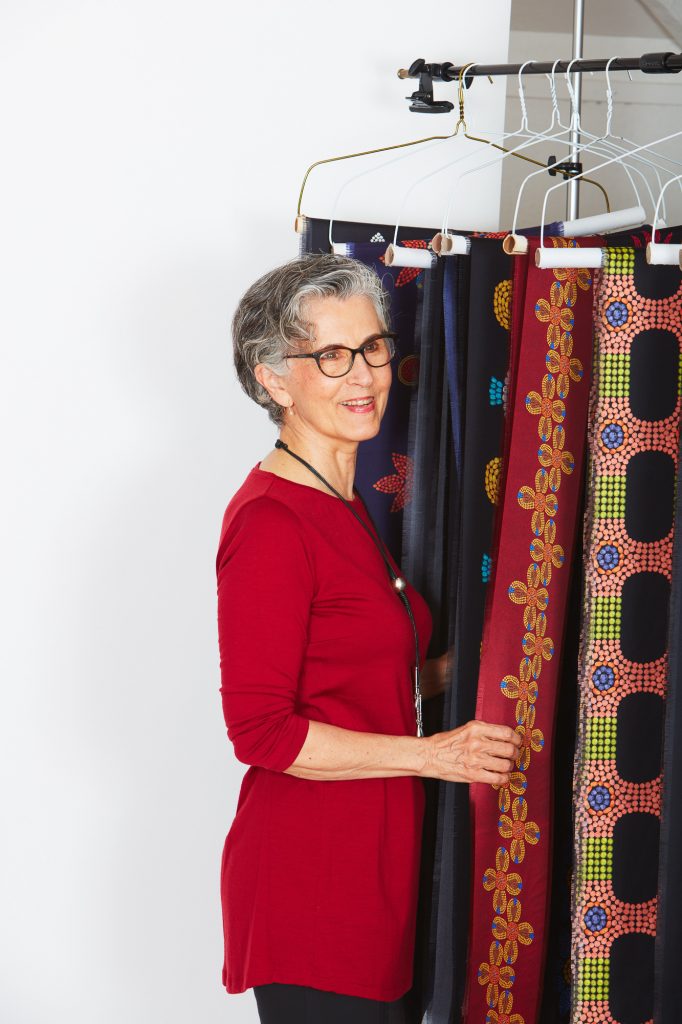 A portrait of Daphne Boyer. She is posed with the ribbons from her work "Birthing Tent" and is wearing a red dress with long sleeves over dark pants, glasses, and a necklace. She smiles as she looks away from the camera.